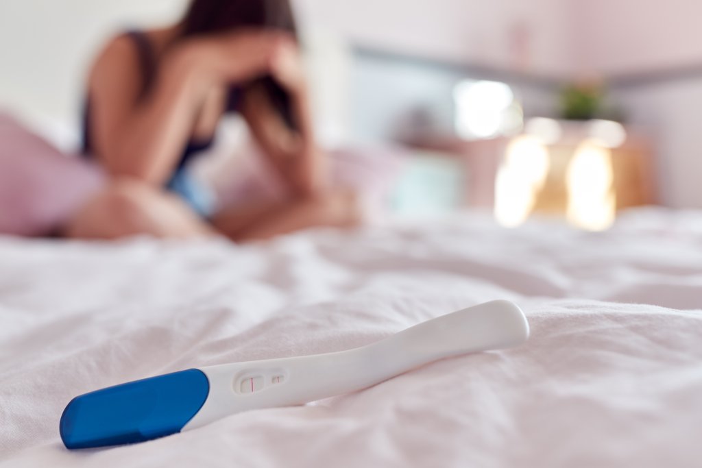 Teenage Girl Sitting On Bed With Head In Hands And Positive Pregnancy Test In Foreground