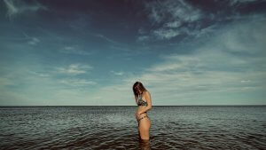 A woman, three months pregnant, strokes her belly while bathing in the sea. Sky it’s cloudy.