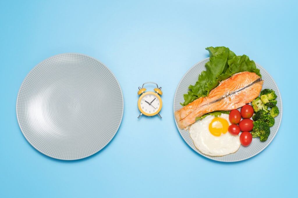 Plates empty and with food and an alarm clock on blue background, concept of intermittent fasting.